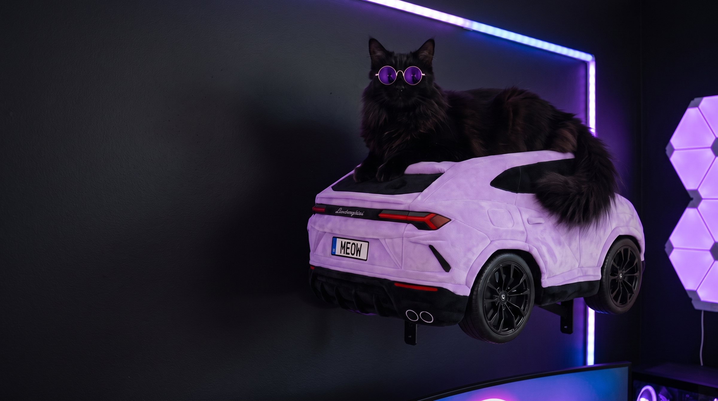 Lilac plush SUV cat bed mounted on a dark gamer-room wall, a black cat in sunglasses on its roof